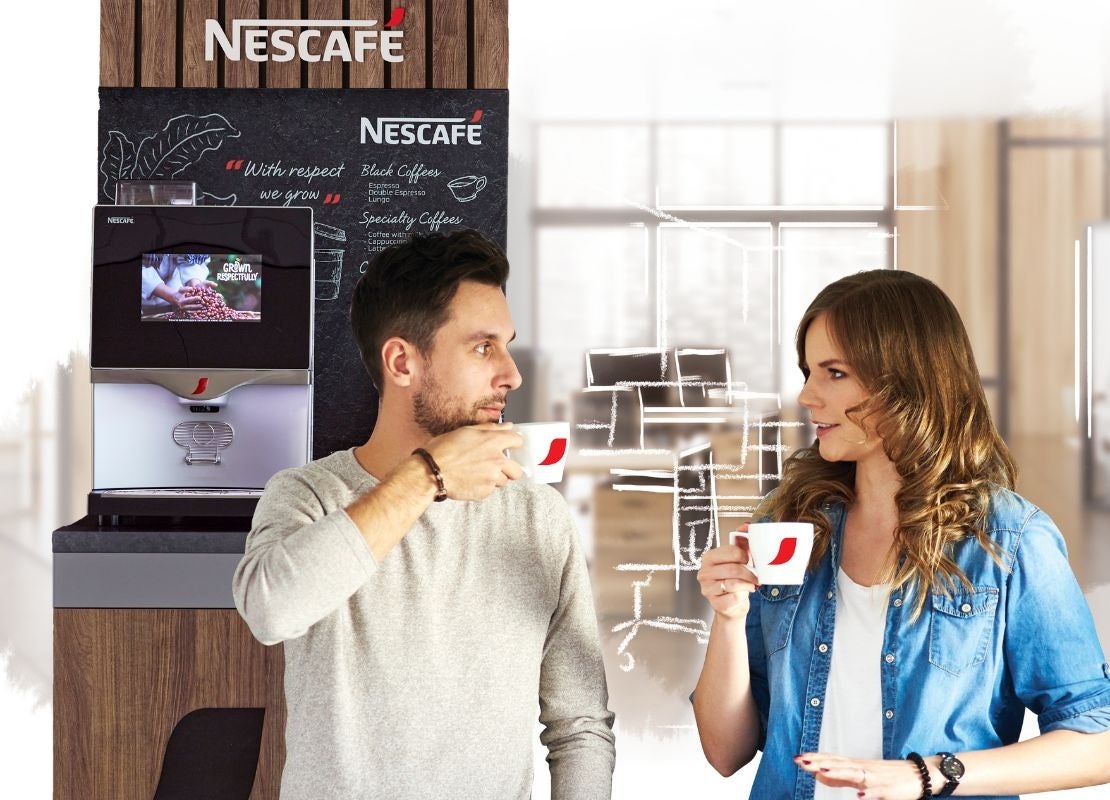 Dos compañeros de trabajo disfrutan de una taza de café en la oficina, al fondo una máquina NESCAFÉ®