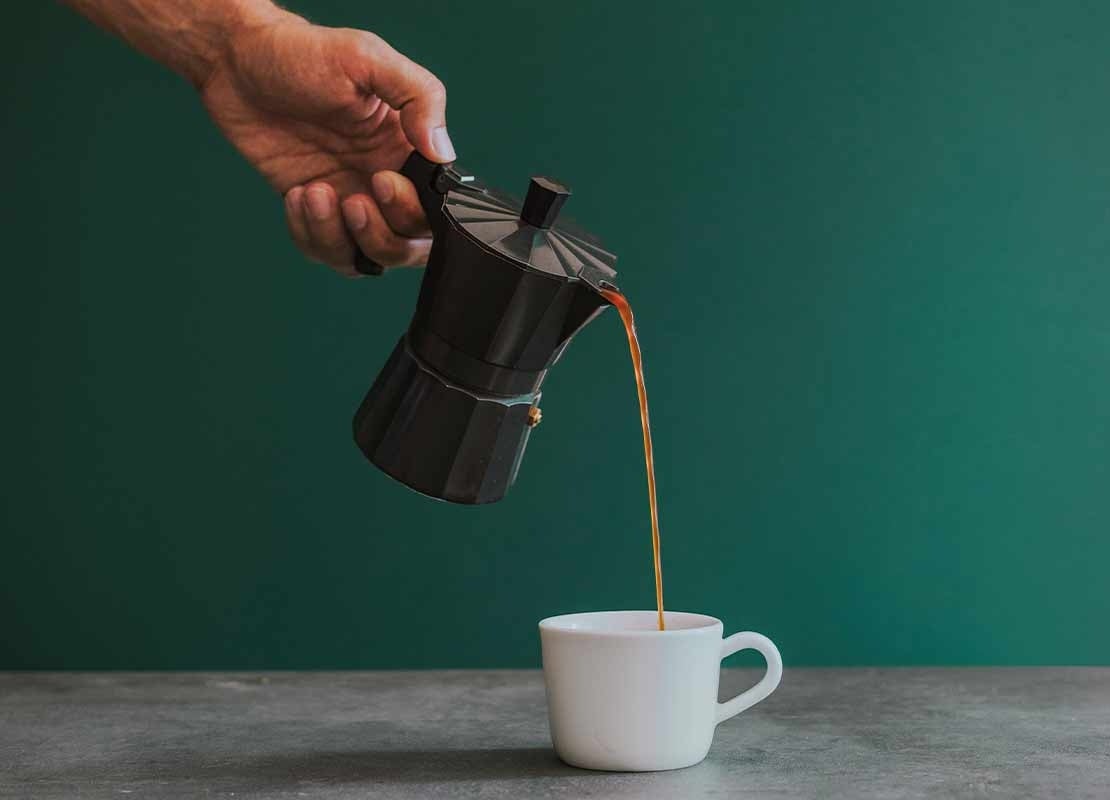 Mano sostiene una cafetera que vierte a una taza café preparado por método de percolación