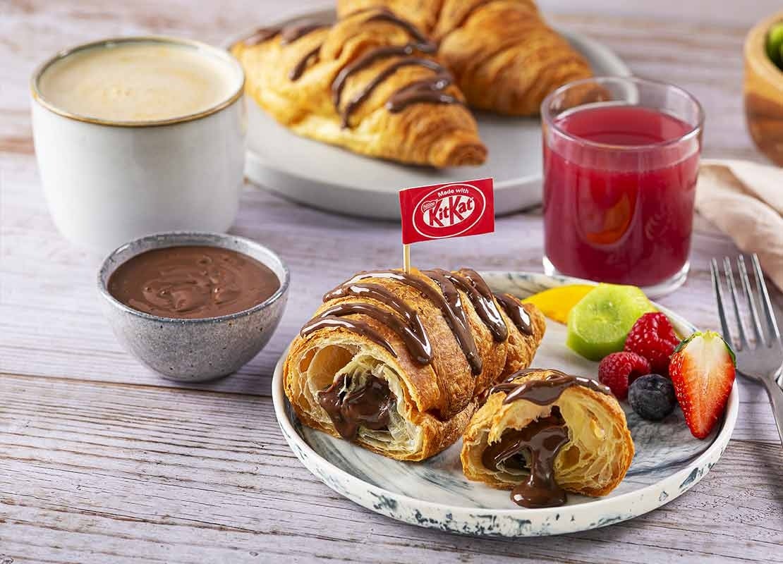 Desayuno servido con croissant con KITKAT®, salsa de chocolate, jugo, frutas y cappuccino NESCAFÉ®