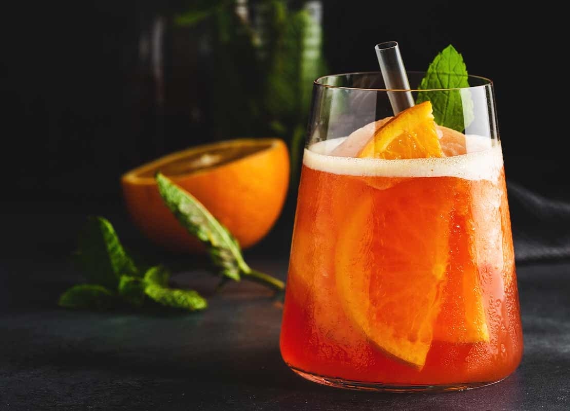 Vaso de cristal con cóctel sin alcohol de naranja y hierbabuena, al fondo la fruta y las hierbas aromáticas