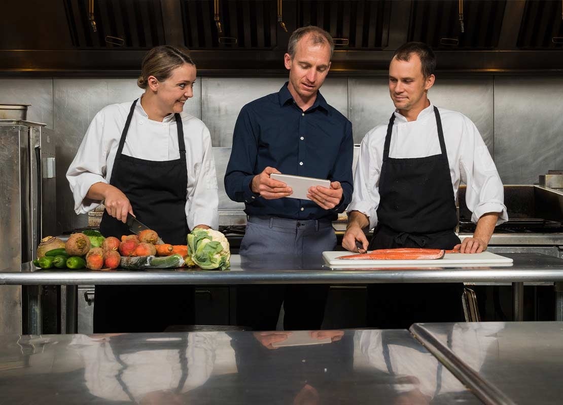 Dueño de restaurante y dos chefs observan una tablet, al frente una mesa con alimentos en una cocina comercial