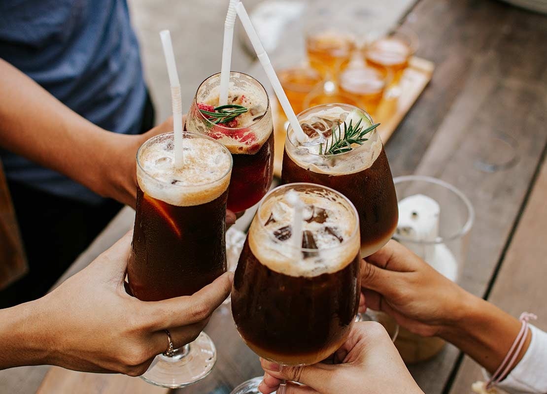Manos sostienen 4 copas con bebidas craft, hielo y pitillos, las cuales chocan para brindar