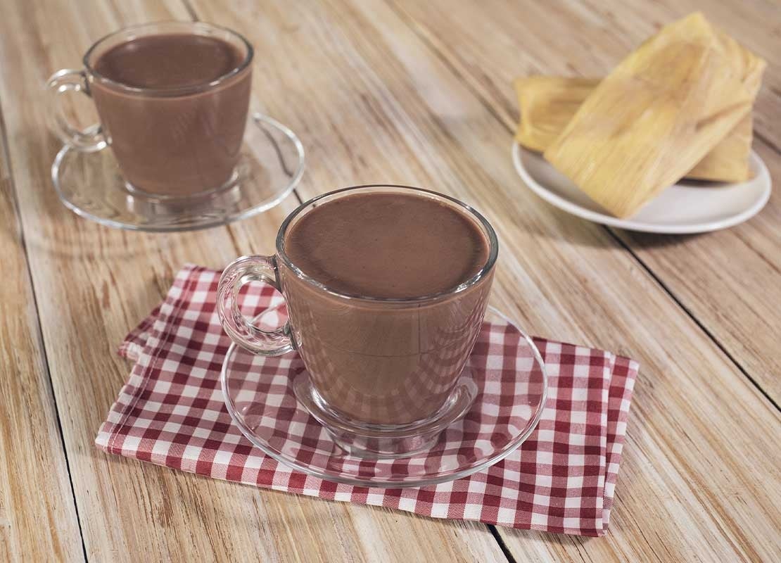 Dos tazas de cristal y sus platos con chocolate caliente, al lado, tamales de maíz