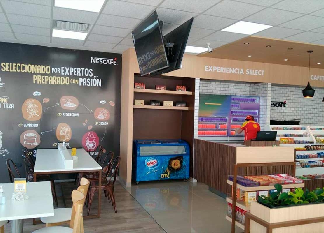 Cafetería de una estación Shell con estanterías, productos, mesas y un banner de NESCAFÉ® en la pared