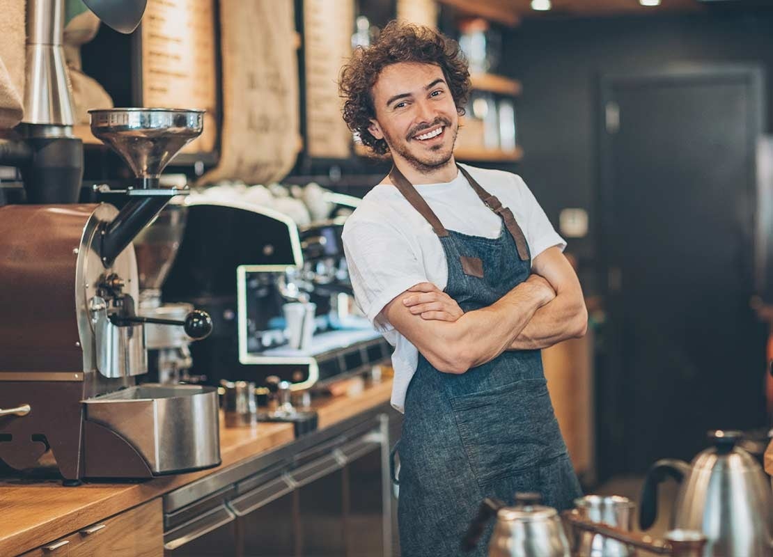 Joven barista con un delantal azul y cruzado de brazos sonríe en la cocina de una cafetería