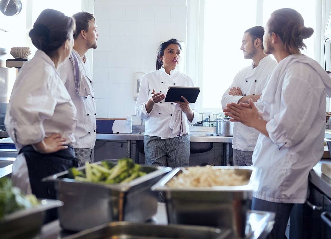 En una cocina comercial, chef sostiene una tablet y explica a 4 cocineros para capacitarlos