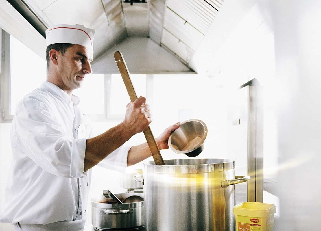 En una cocina pequeña, chef mezcla una olla con una cuchara de madera mientras agrega un sazonador
