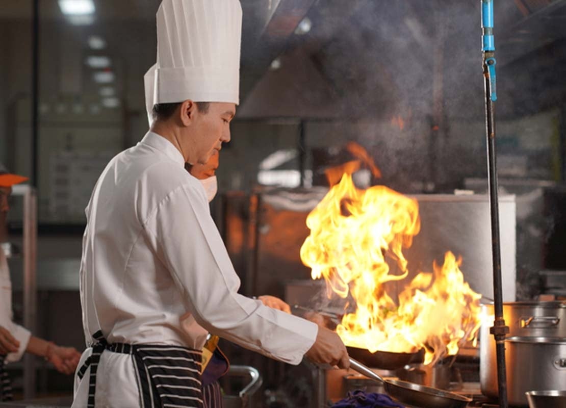 Chefs con gorra y delantal sostienen sartenes ardiendo sobre la estufa de una cocina comercial