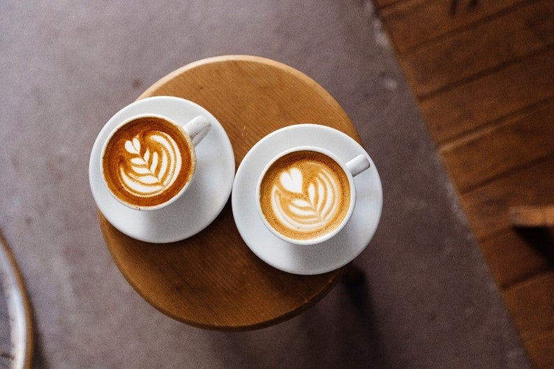 Dos tazas de café latte con arte en espuma, colocadas sobre una mesa de madera en un entorno acogedor.