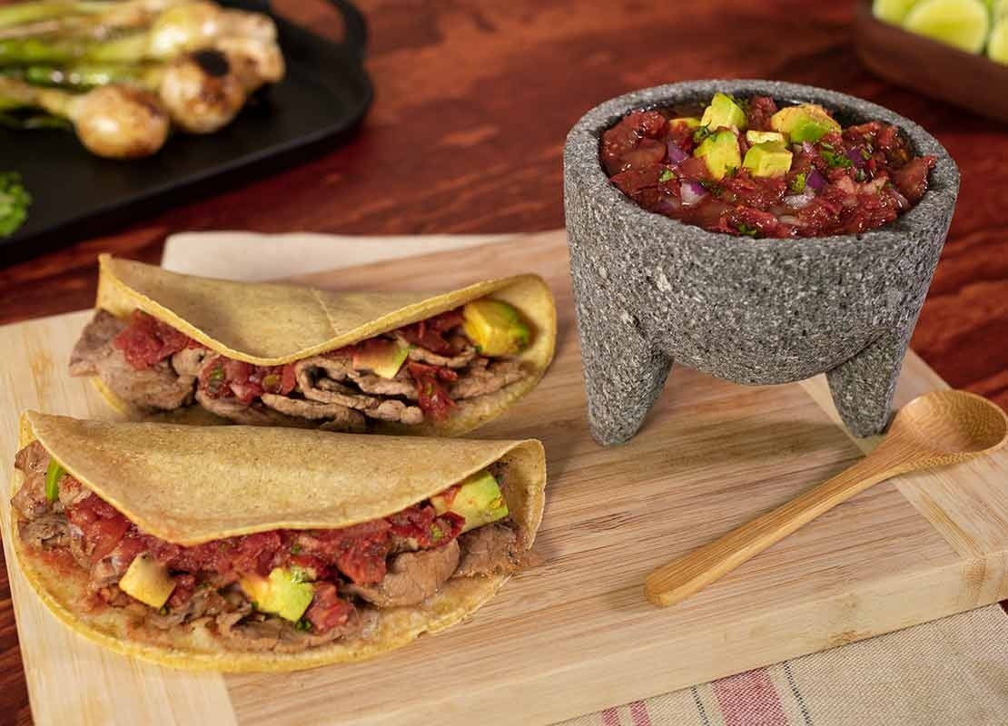 Tabla con dos fajitas de carne, salsa en molcajete, cuchara de madera, al fondo bandeja con cebollas
