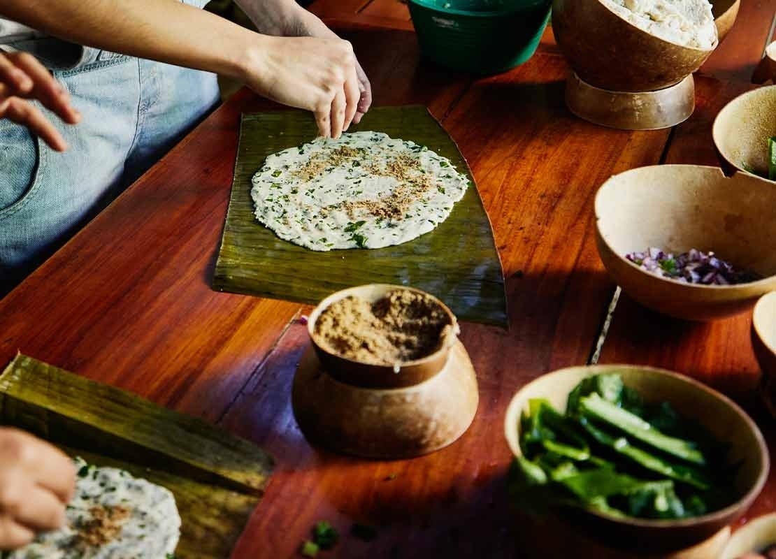 Mesa con cuencos rodeada por personas que envuelven alimentos autóctonos en hojas de plátano