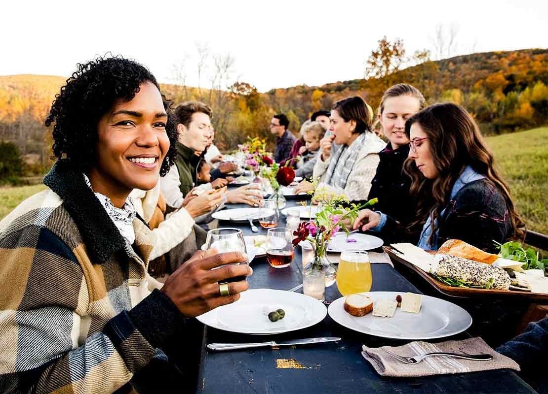 Grupo de amigos comen al aire libre sentados en una mesa de madera, rectangular y larga