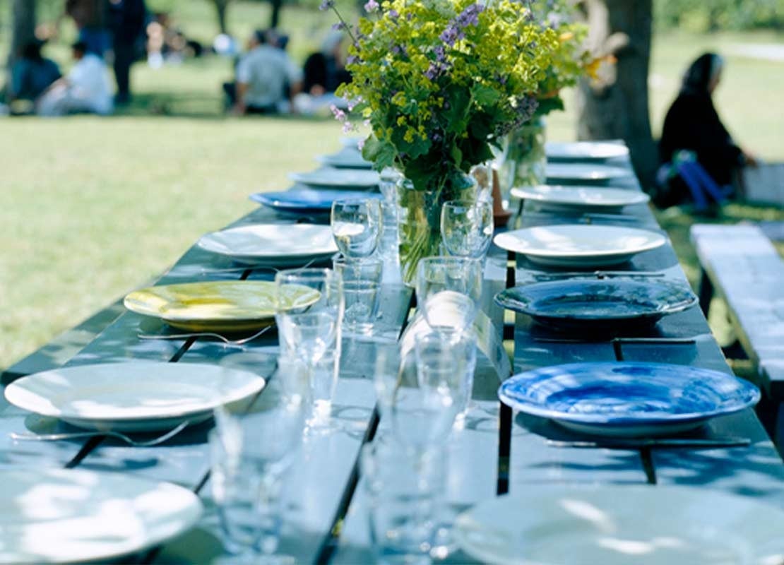 Mesa rectangular con hileras de platos y vasos dispuesta para servir comida al aire libre