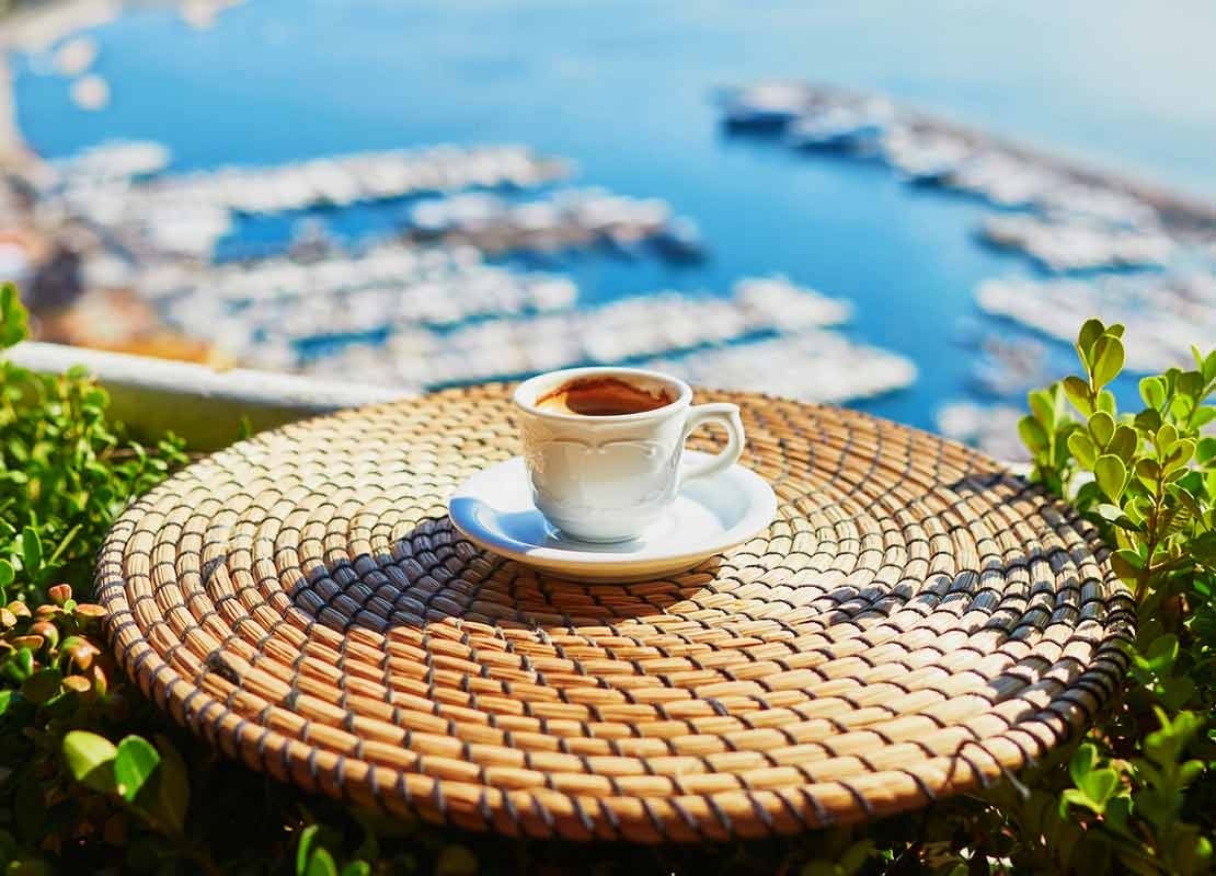 Taza de café servida sobre una mesa en un balcón, al fondo orilla del mar con casas y muelles