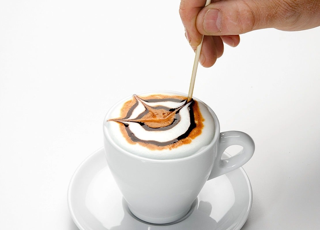 Mano decorando un café con arte latte usando un palillo sobre espuma y jarabe