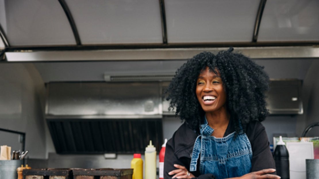 Persona sonriente frente a un food truck con insumos y estación de trabajo al aire libre.