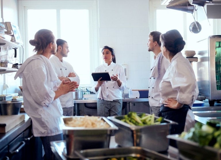 Equipo de cocina escuchando a chef con tablet durante reunión en cocina