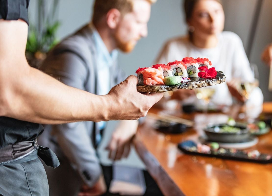 Mesero sirviendo plato de sushi a una mesa con personas comiendo comida de Asia Oriental con palillos