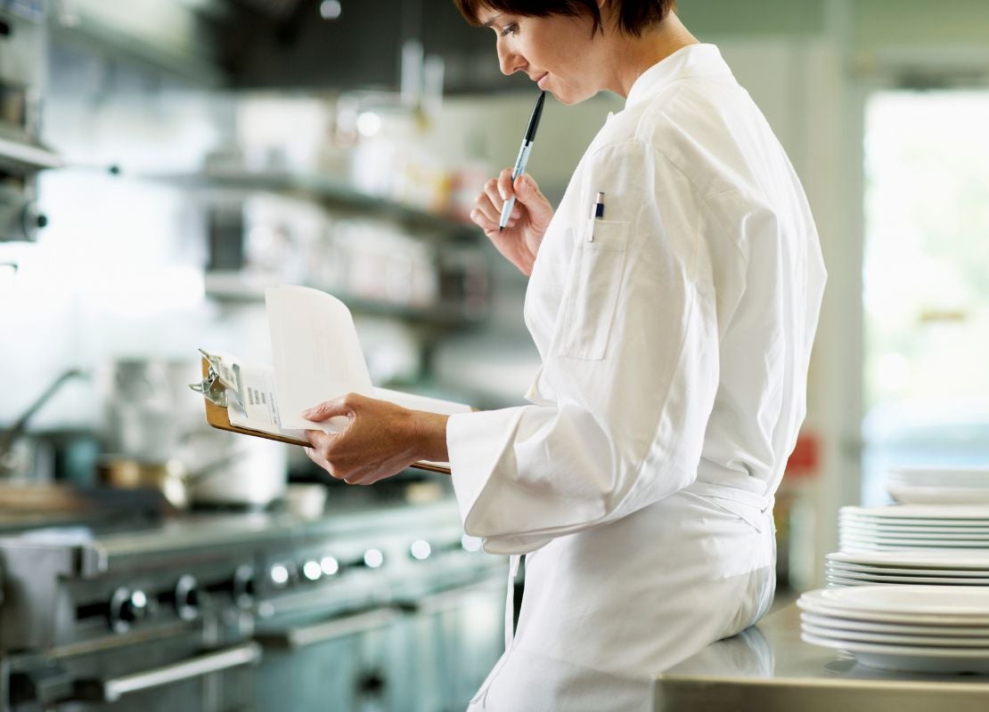 Una chef con uniforme revisa una planilla mientras sostiene un bolígrafo en una cocina industrial