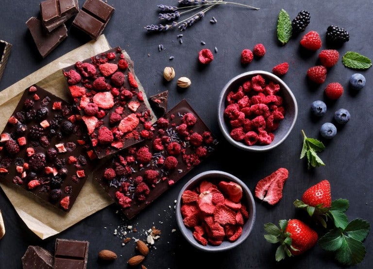 Barras de chocolate amargo con frambuesas y moras deshidratadas, junto a cuencos de frutos liofilizados.