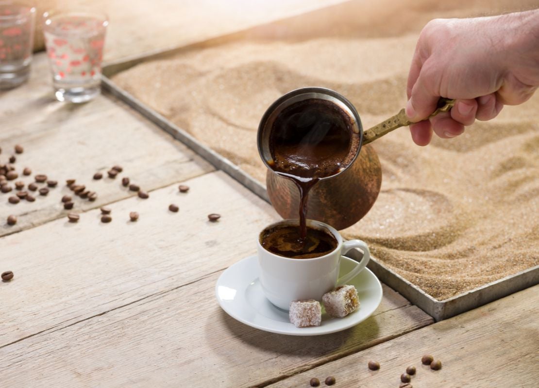 Café turco servido desde cezve (ibrik) en una taza sobre una mesa con calentador de arena
