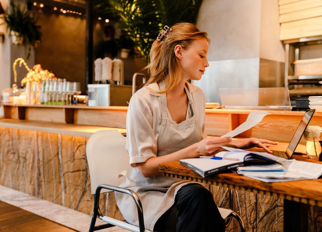 Propietaria de restaurante revisa documentos físicos junto a su laptop en la barra del local.