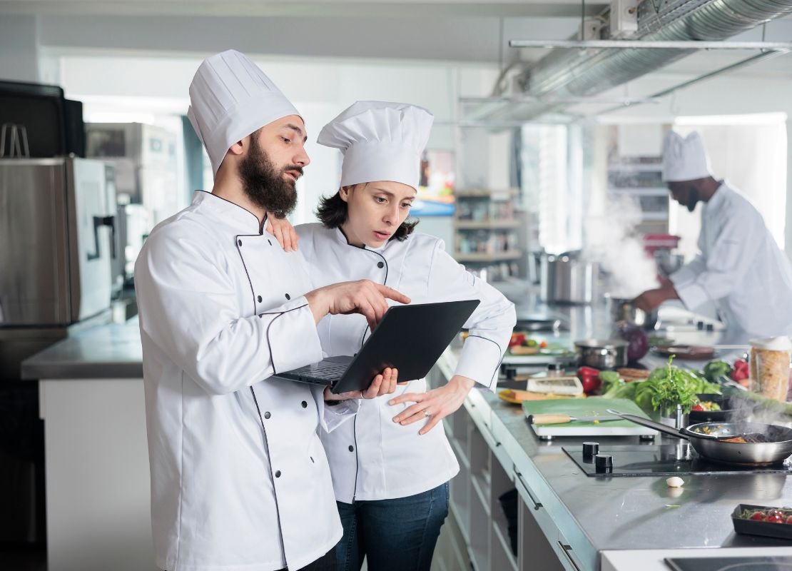 Dos chefs uniformados revisan una computadora portátil dentro de una cocina profesional.