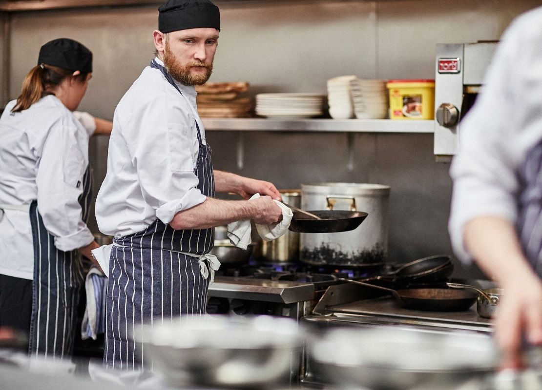 Chef cocinando con sartén frente a estufa en cocina profesional con otros cocineros