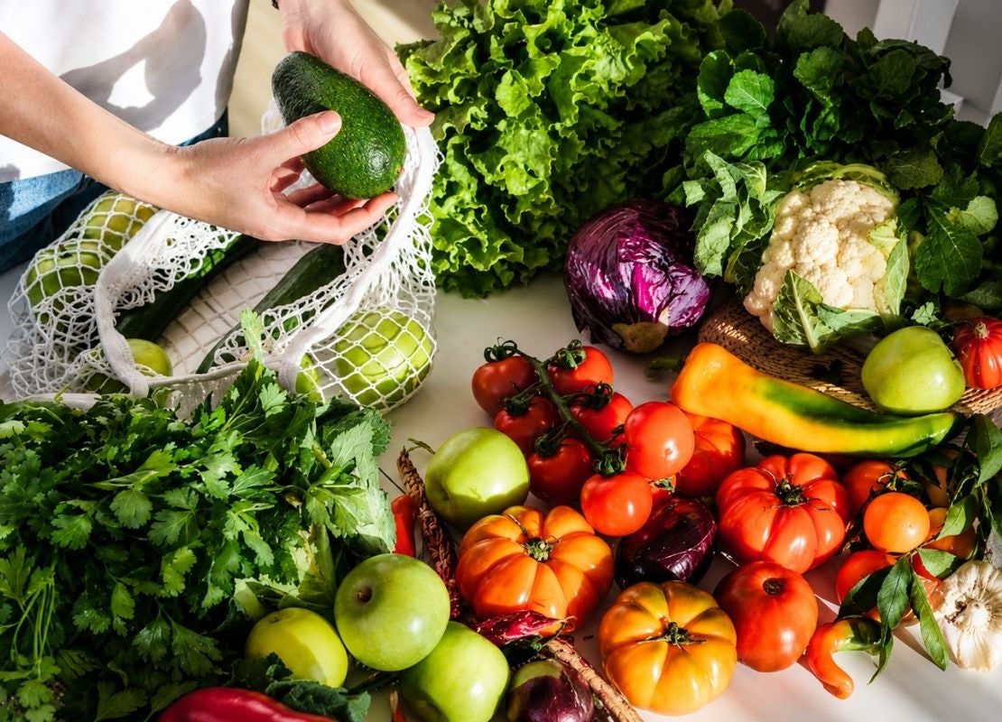 Manos guardando un aguacate en una bolsa de malla junto a tomates y vegetales frescos variados.