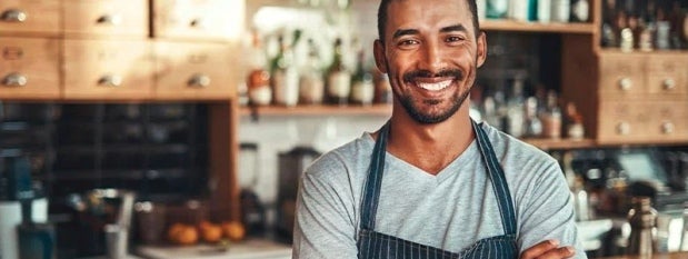 Un barista cruzado de brazos sonríe y a su espalda está la cocina de una cafetería equipada con ingredientes e instrumentos