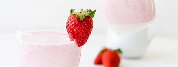 Batido en un vaso y en el borde una fresa anclada como elemento decorativo, al fondo una taza blanca