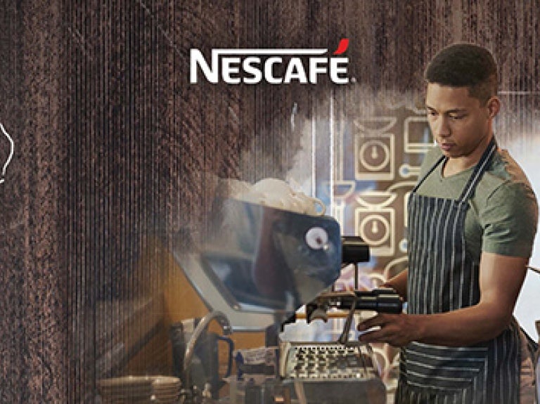 Joven preparando una taza de café en una Máquina Nescafé mientas es observado y evaluado por una mujer joven