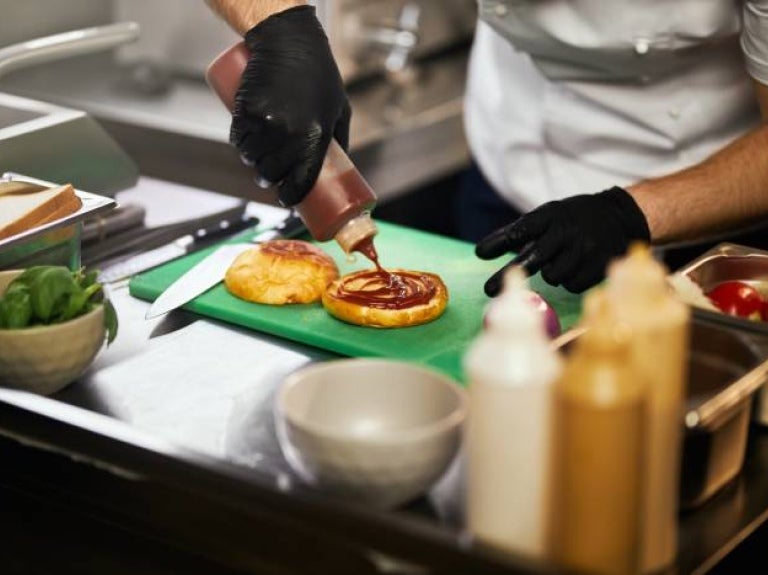 Chef con guantes negros añadiendo salsa sobre panes de hamburguesa