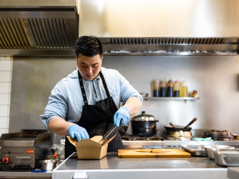 En una cocina comercial, chef empaca una preparación en una caja para ser enviada como entrega a domicilio