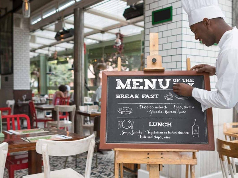 Chef escribe un menú de restaurante sobre una pizarra frente a su negocio que es espacioso y colorido