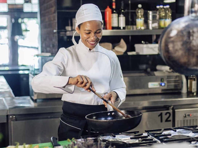 Una chef prepara comida en una sartén, rodeada de un equipamiento de cocina ajustado a las necesidades de un restaurante