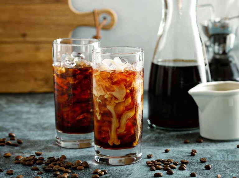 Dos vasos de café frío con hielo y leche sobre una mesa junto con granos de café tostados y una jarra con la bebida