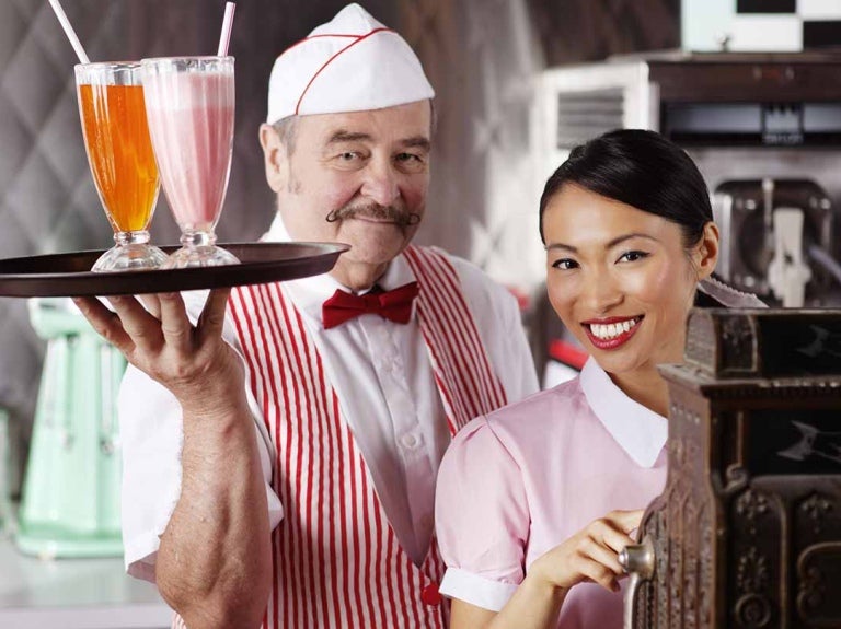 En restaurante temático de los 50, un mesero sostiene una bandeja con jugos y otra opera la caja registradora