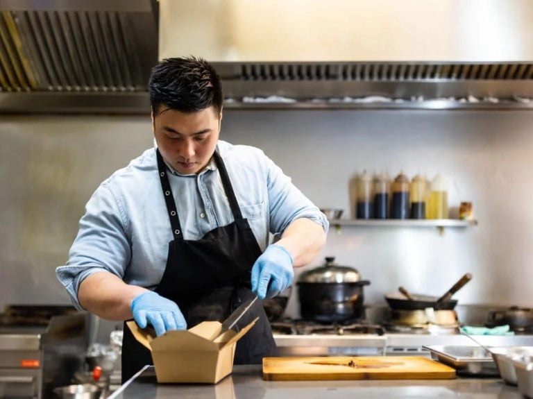 En una cocina comercial, chef empaca una preparación en una caja para ser enviada como entrega a domicilio