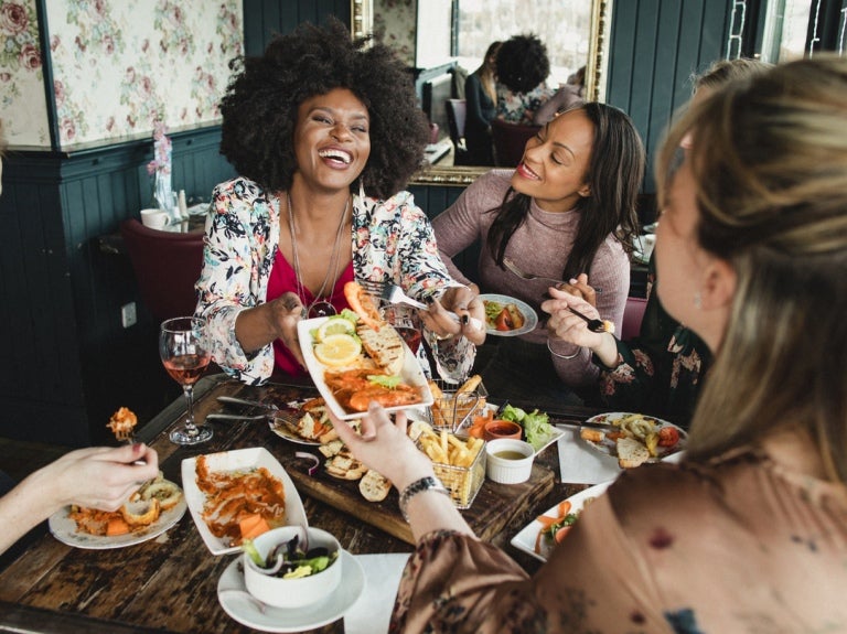 Grupo de 5 amigas sonrientes conmemoran el Día Internacional de la Mujer al compartir un almuerzo especial en un restaurante