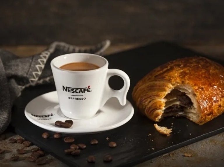 Una taza de espresso NESCAFÉ® y un croissant de chocolate mordido rodeados de granos de café