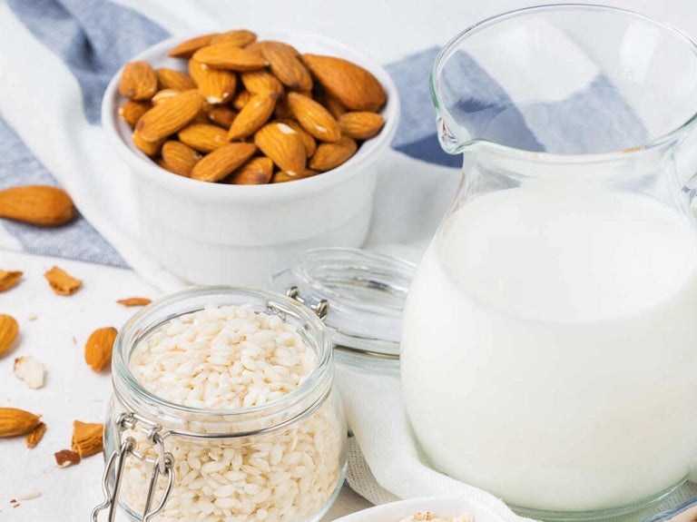 Sobre una mesa una jarra con una bebida vegetal y a su alrededor recipientes con avena, arroz, almendras y garbanzos