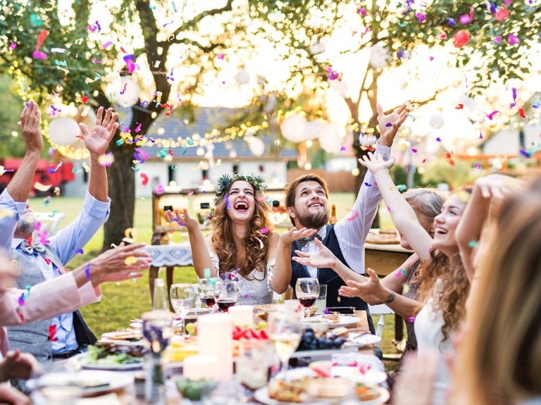 Recién casados y sus invitados vitorean mientras están sentados a la mesa al aire libre con platillos servidos como picnic