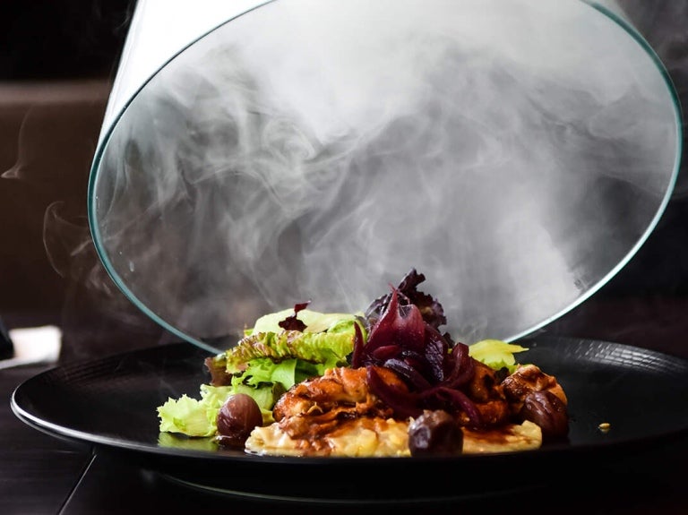 Sobre una mesa de madera se destapa un plato de comida gourmet envuelta en humo