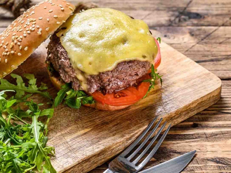 Sobre una tabla de cortar, una hamburguesa tradicional y al lado un par de cubiertos