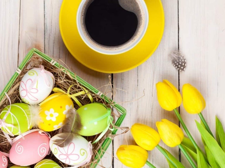 Sobre una mesa de madera 5 rosas amarillas, una taza de café y una canasta con dulces de pascua como huevos decorados