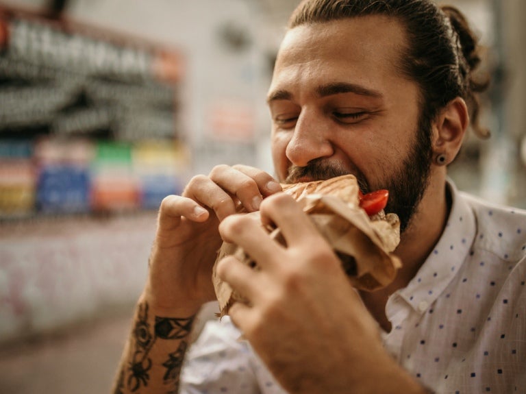 Vista frontal de un hombre que come una arepa rellena, la cual sostiene con sus dos manos, fuera de un negocio de street food