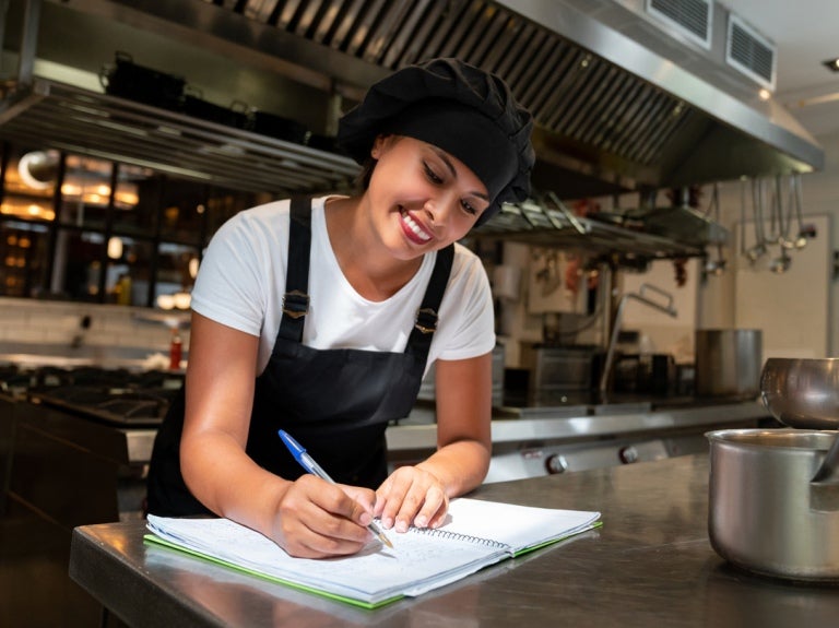 En una cocina comercial, joven estudiante del programa YOCUTA toma notas en su libreta sobre