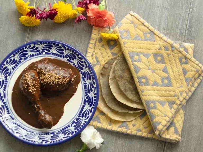 Plato con dos piezas de pollo cubiertas de la salsa mole, al lado 4 tortillas de maíz en un mantel
