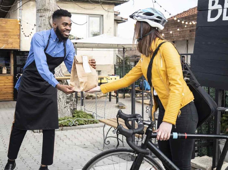Empleado de restaurante entrega a una clienta que va en bicicleta un pedido a través de take away